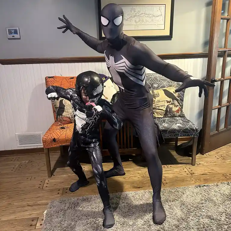 Two people in superhero costumes pose indoors: one in a black and white Spider-Man suit with arms outstretched, and a…