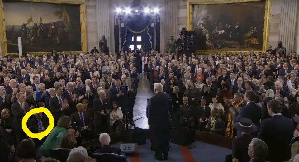 A wide shot of a formal event in a grand hall with many people in suits applauding. A man in the audience, circled in…