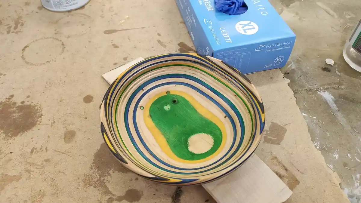 A handcrafted wooden bowl with concentric painted rings in blue, green, and yellow sits on a workbench. The bowl's interior…