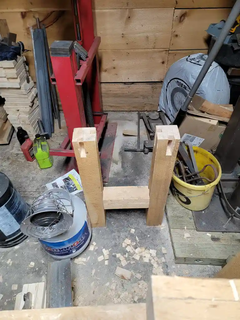 A workshop scene shows two wooden chair legs with a horizontal wooden seat piece between them, forming a chair frame on a…