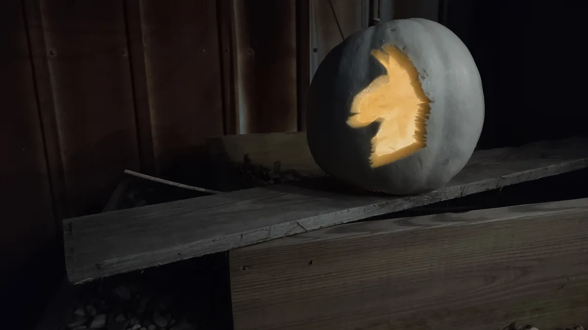 A white pumpkin sits on a wooden plank at night, carved with a horse's head silhouette that glows from within. The text…