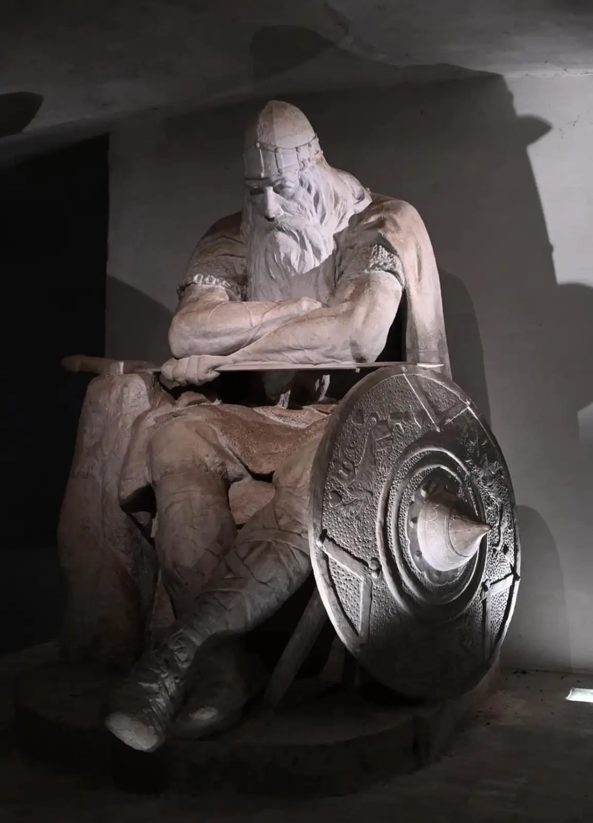 A large stone statue of a bearded Viking warrior sits in a throne-like chair, arms crossed over his chest, with a detailed…