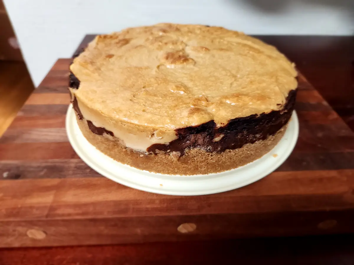 A round dessert with a golden-brown, baked top layer sits on a white plate atop a wooden cutting board. The side reveals a…