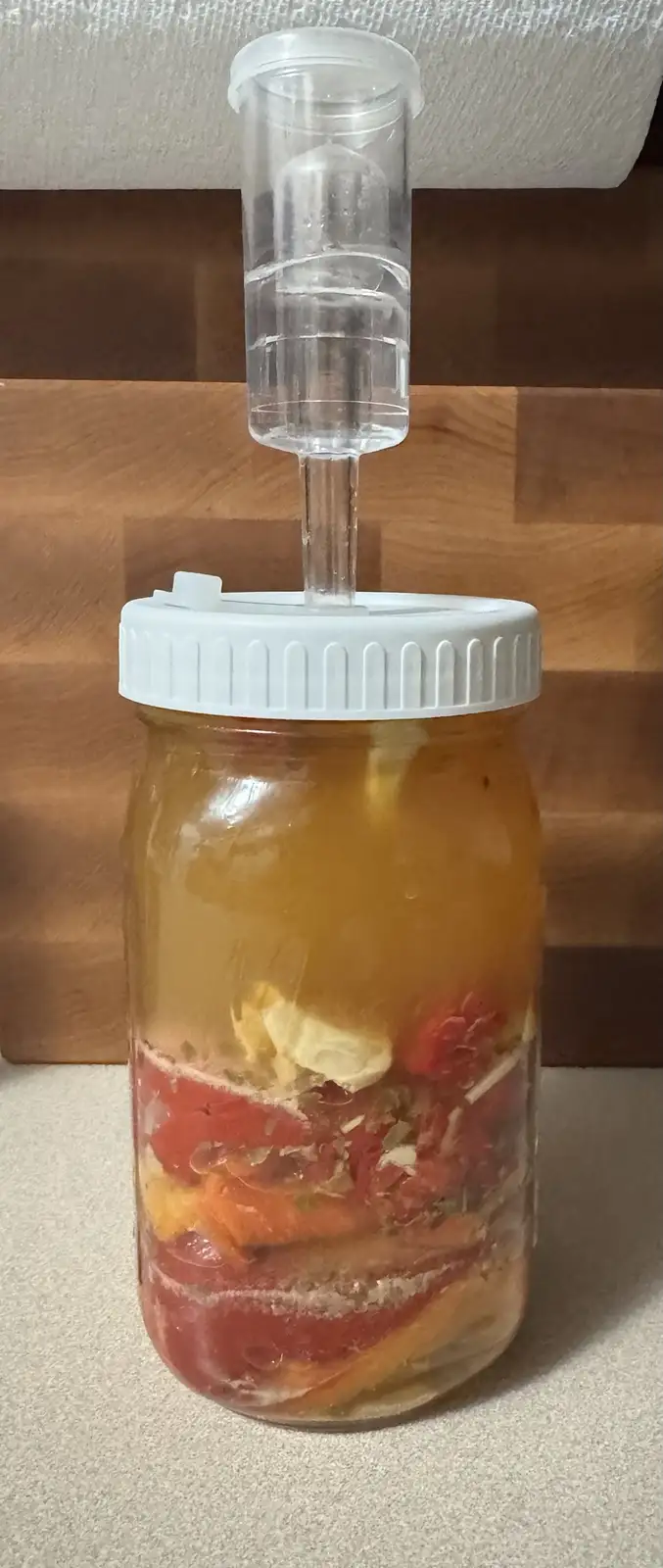 A glass mason jar filled with sliced red and orange peppers, garlic, and brine, topped with a white lid and a clear airlock…