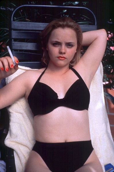 A woman in a black bikini sits on a lounge chair, holding a cigarette, with a towel draped over her shoulder. Her expression…