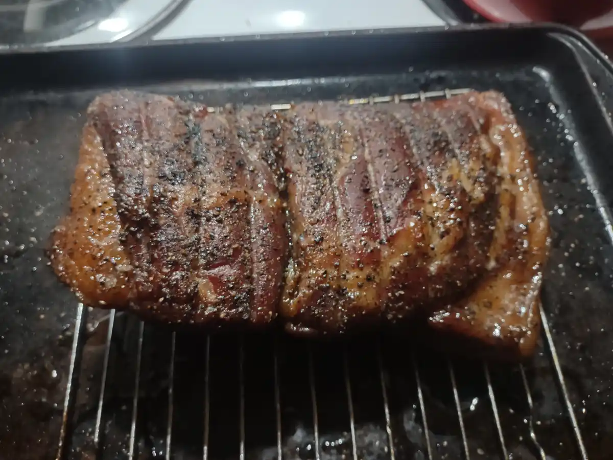 A close-up photo shows a large, seasoned piece of meat, likely ribs or a roast, sizzling on a metal grill rack inside a…