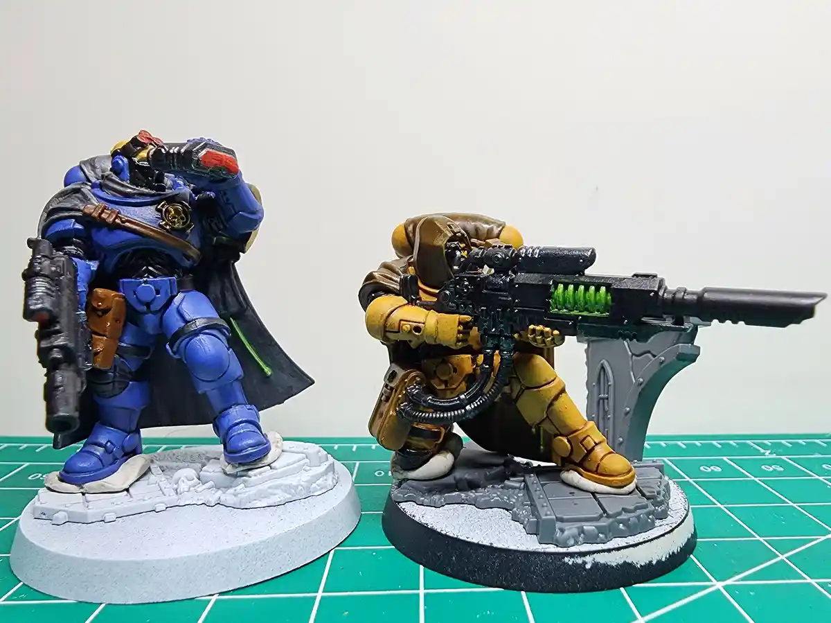 Two painted miniature sci-fi soldiers stand side-by-side on a green grid mat; the blue-armored figure on the left holds a…