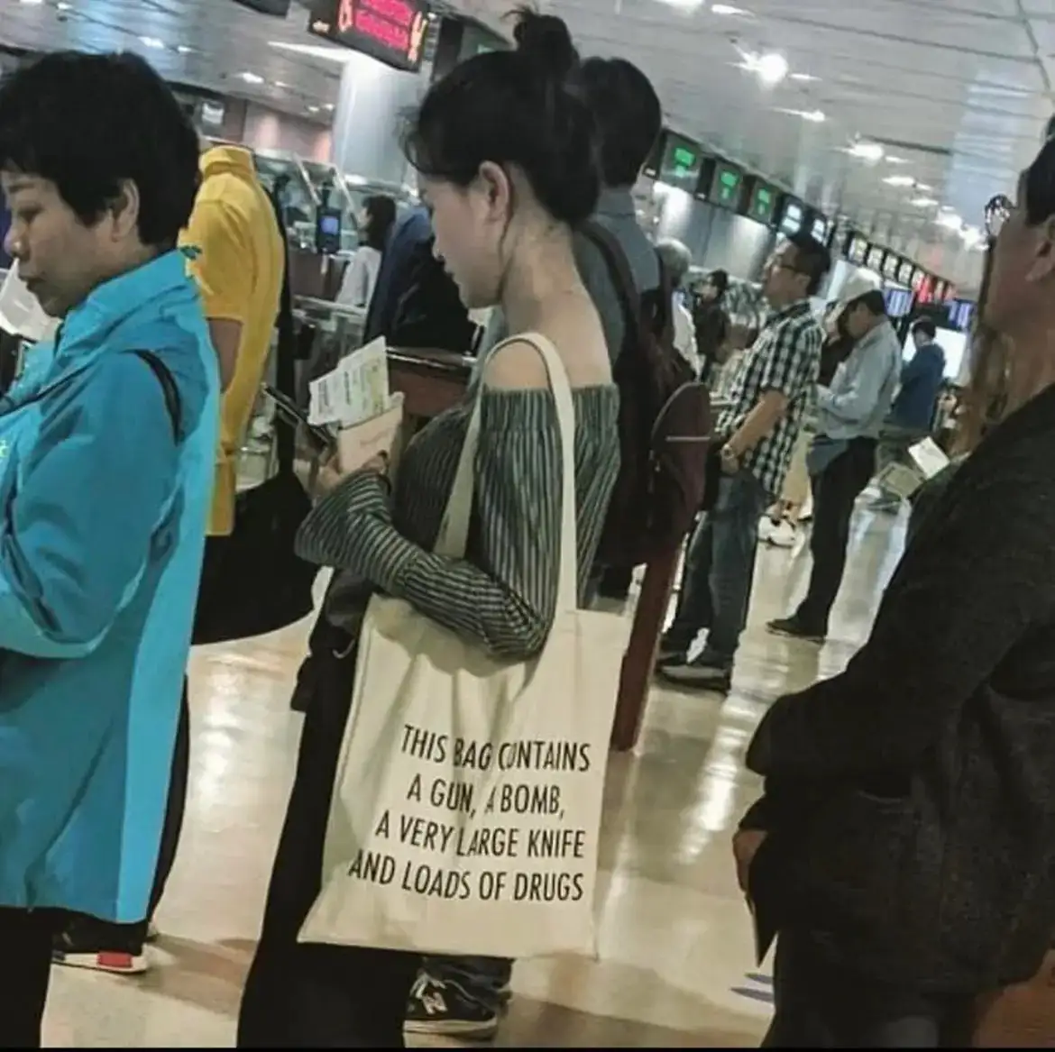A woman stands in an airport queue holding a beige tote bag with bold black text declaring it contains a gun, a bomb, a very…