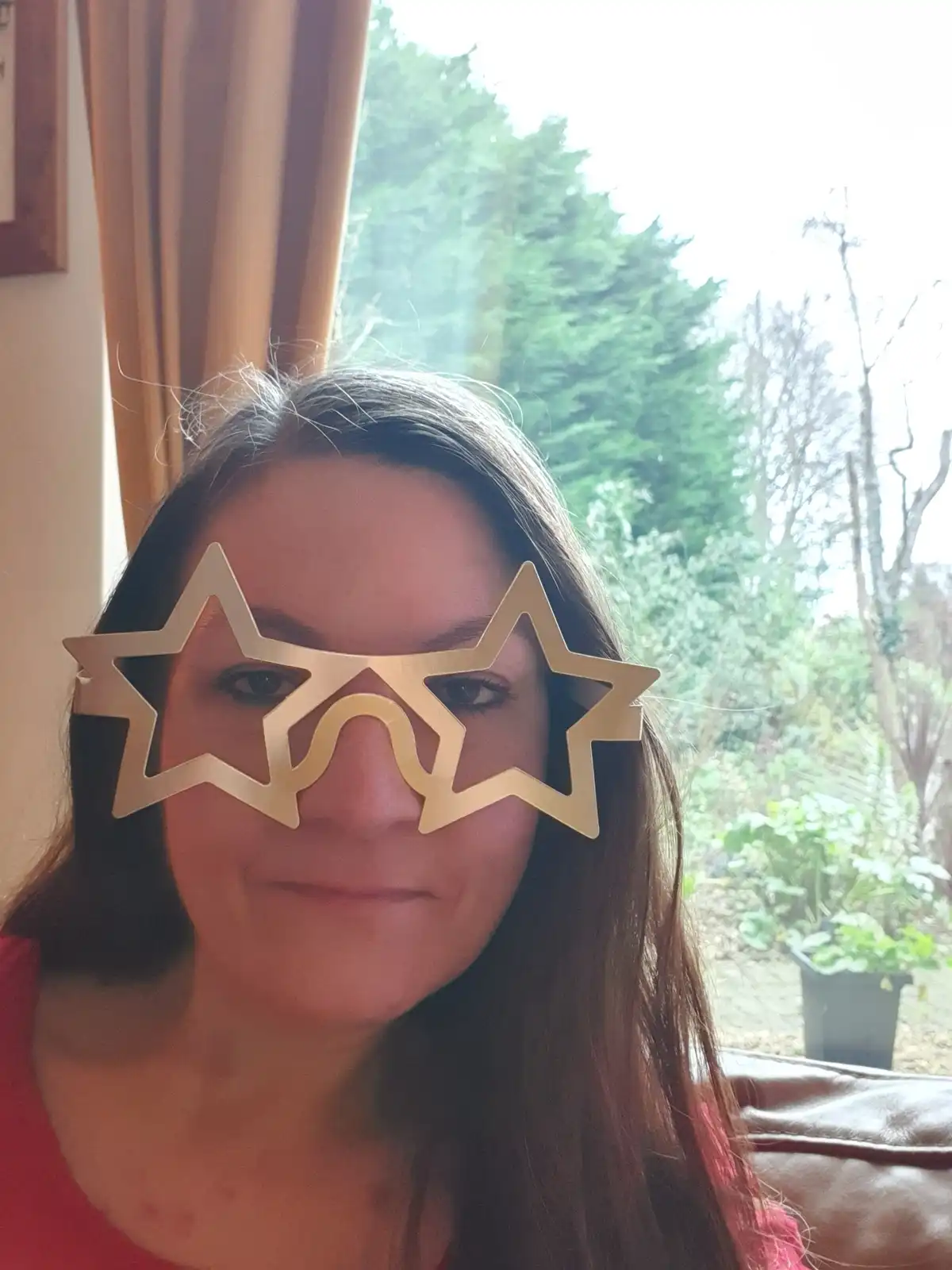 A woman with long dark hair is photographed indoors, wearing large, gold-colored star-shaped glasses that cover her eyes and…