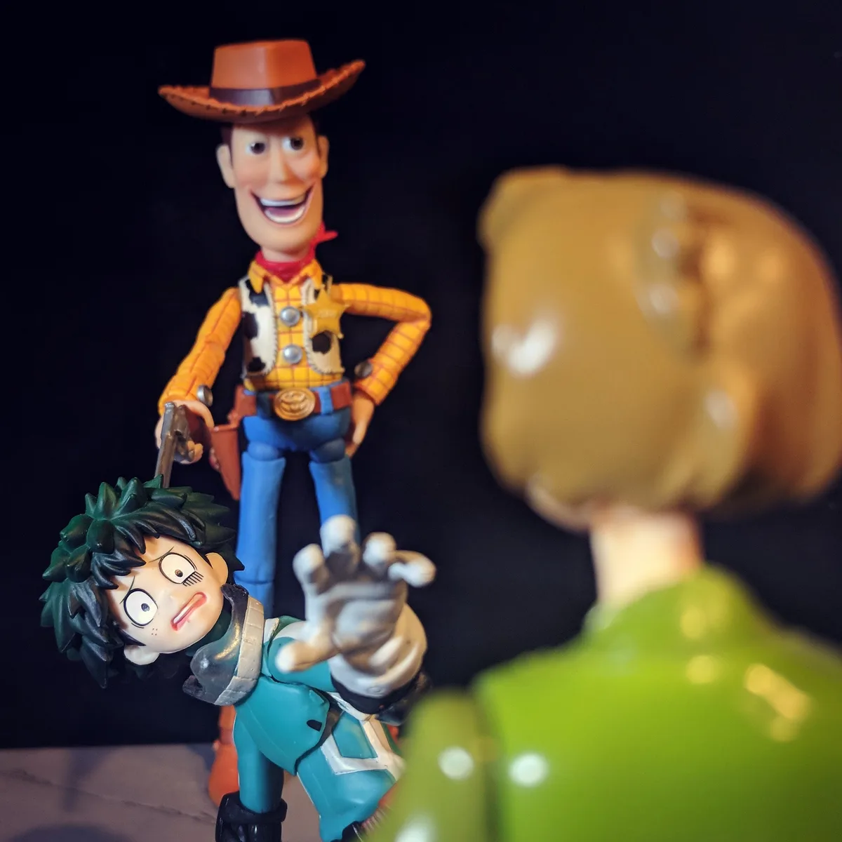 A photo shows three toy figures against a black background. In focus is Woody from Toy Story, smiling and holding a gun…