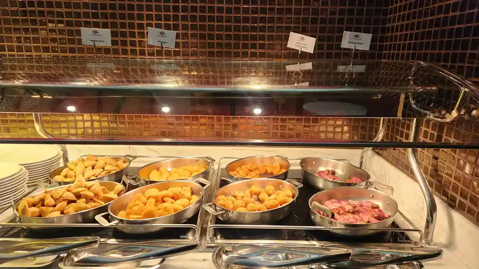 A buffet display with multiple metal pans filled with fried foods like samosas, fish nuggets, cheese balls, and sausage…