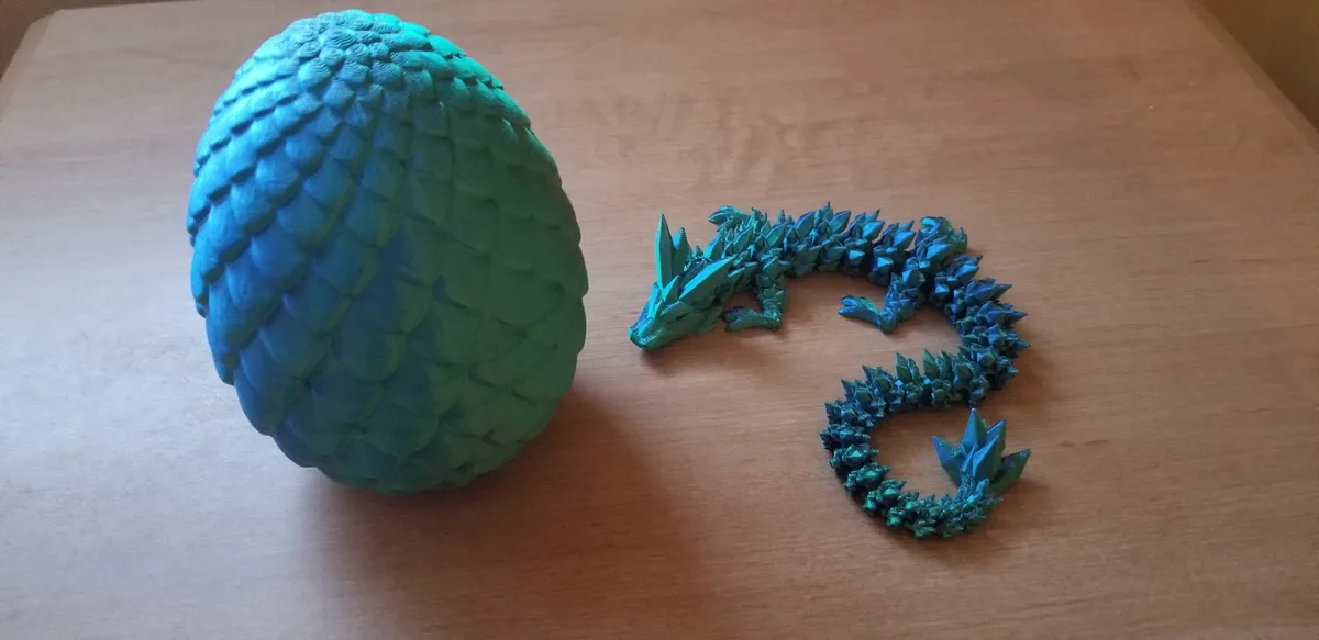 A large, textured blue-green dragon egg sits on a wooden table next to a tiny, similarly colored dragon figurine that is…