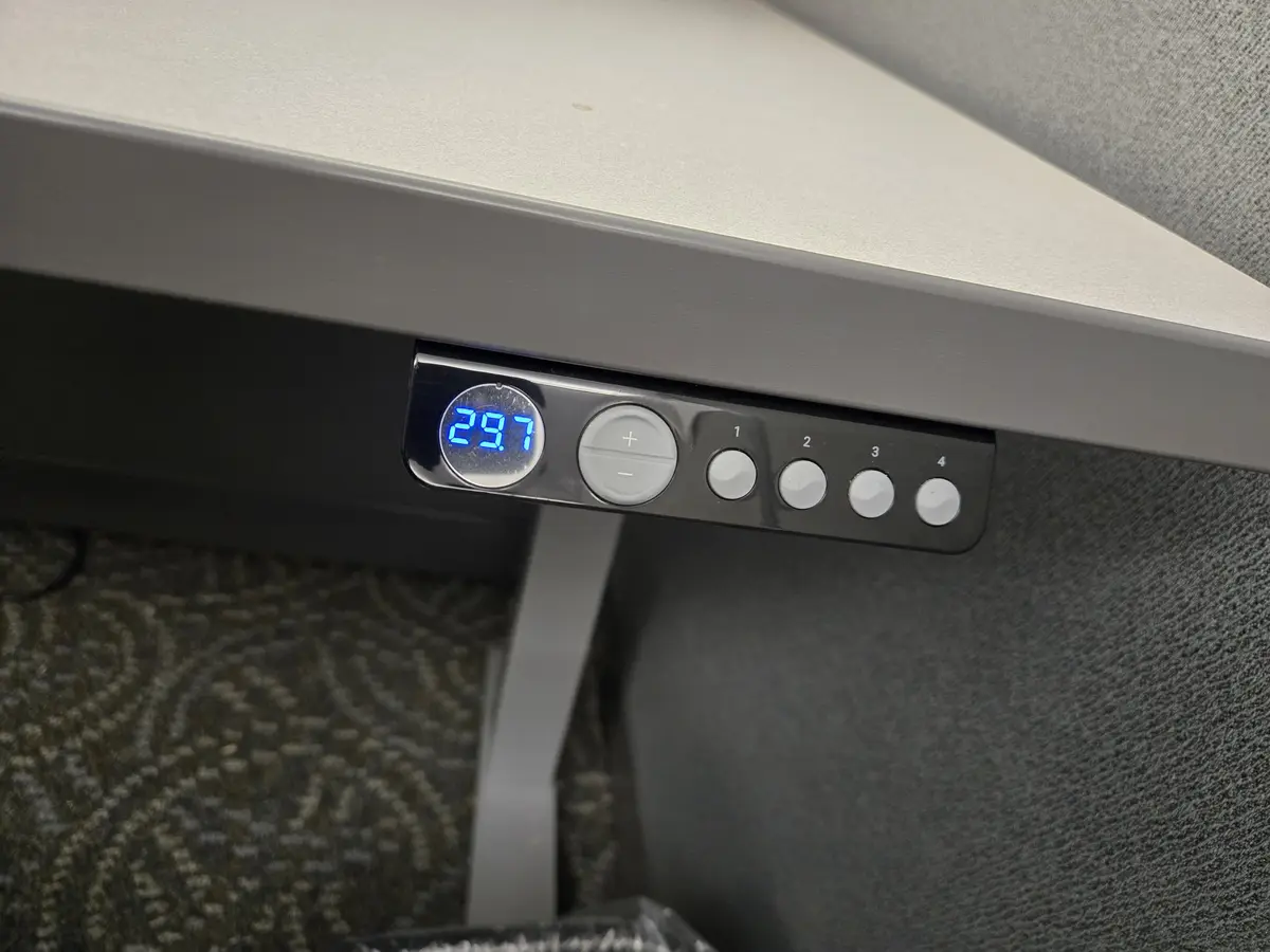 A close-up of an electric desk's control panel shows a digital display reading '29.7' in blue LED numbers, next to…