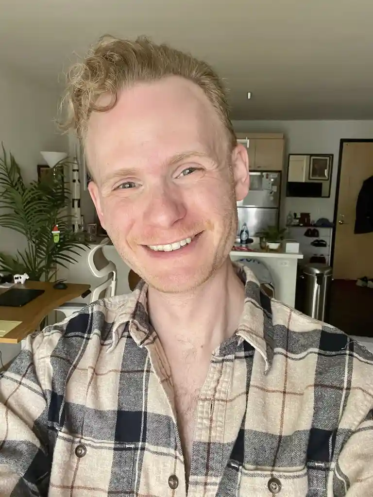 A man with light blonde curly hair and a plaid shirt smiles broadly at the camera in a selfie taken in a home kitchen. The…