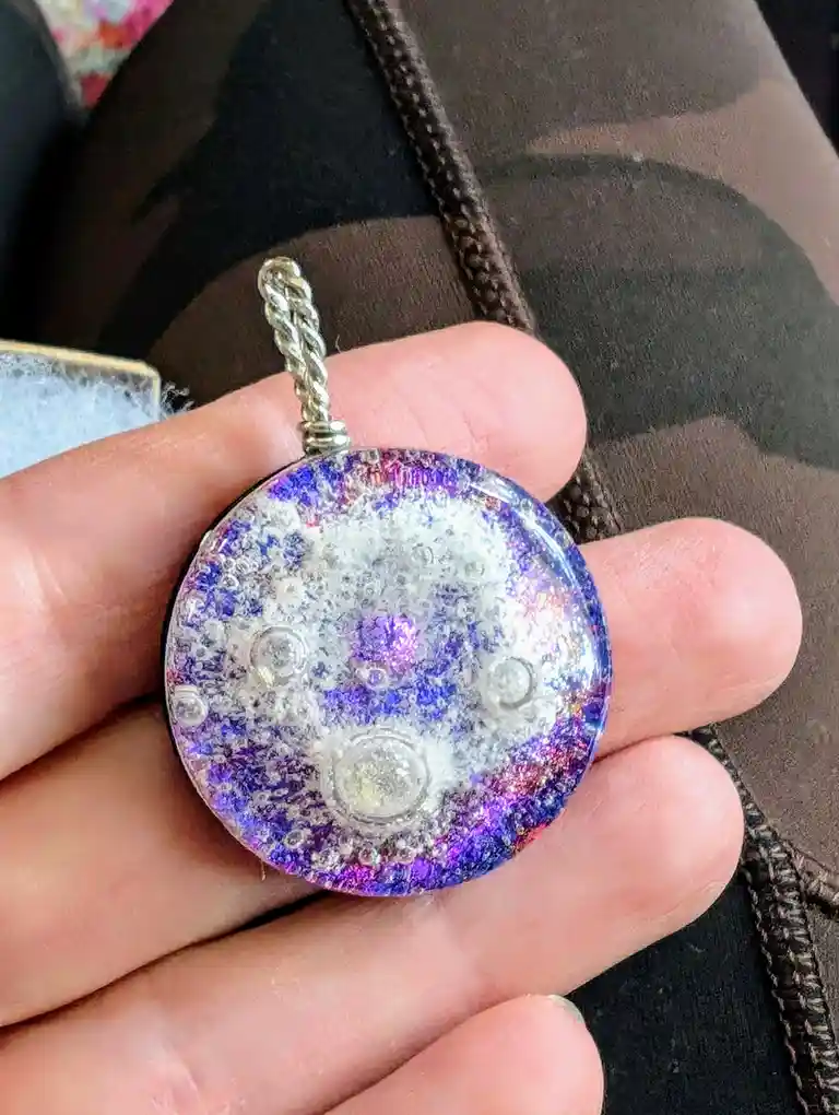 A hand holds a round, iridescent glass pendant with a silver twisted bail, featuring a design resembling the moon's surface…