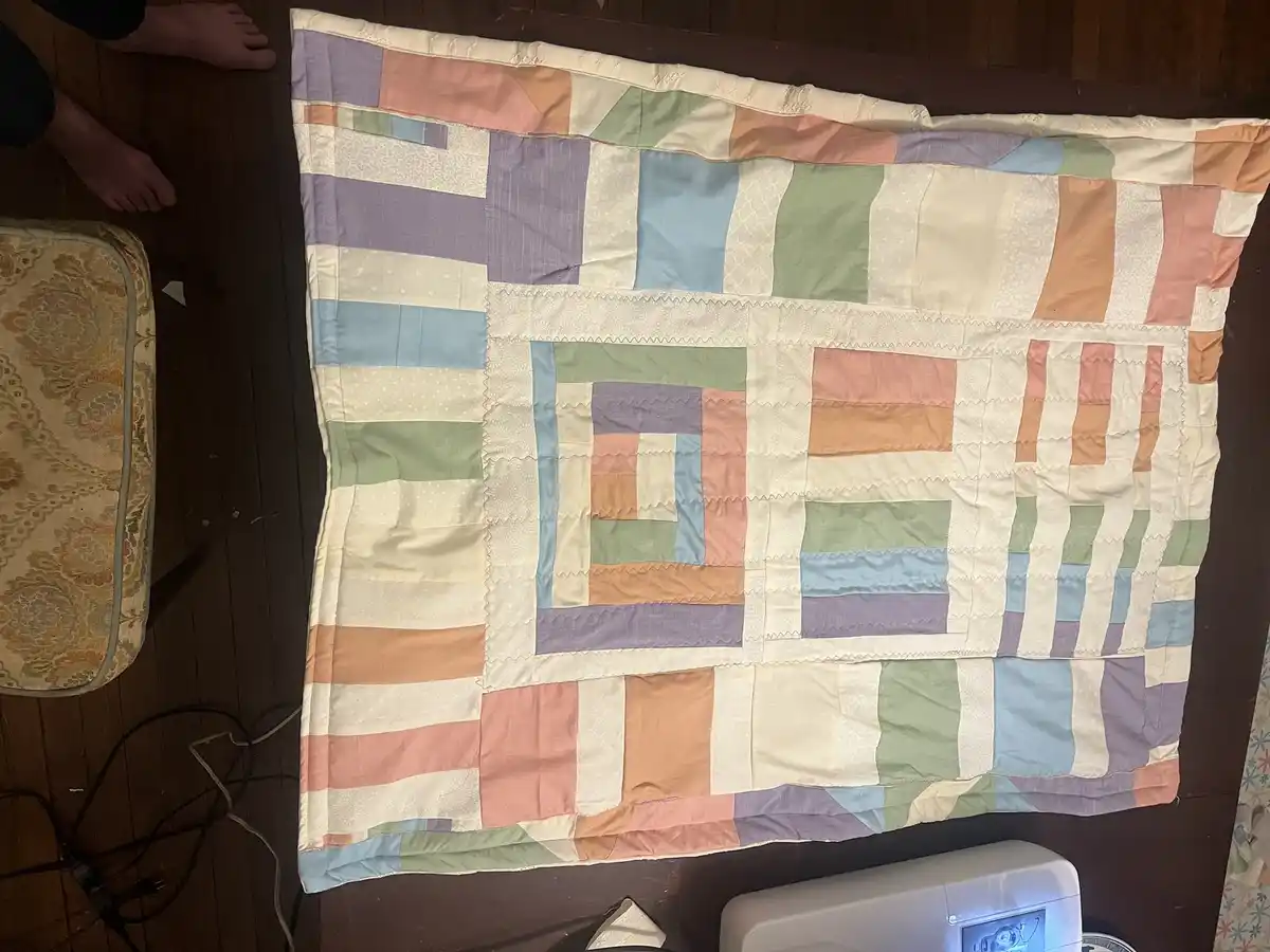 An overhead photo shows a colorful patchwork quilt laid flat on a dark wooden floor, next to a patterned chair and a white…