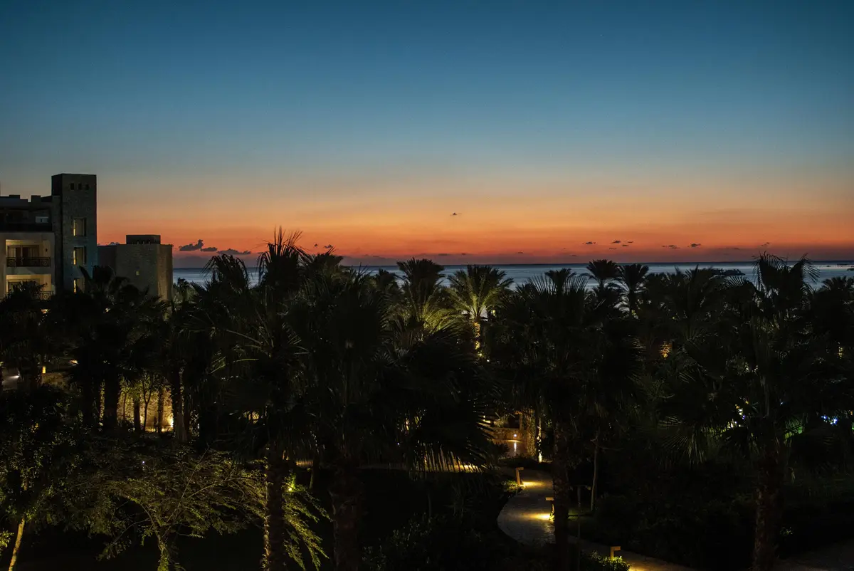 A serene twilight scene shows a resort with palm trees silhouetted against a gradient sky transitioning from orange to deep…