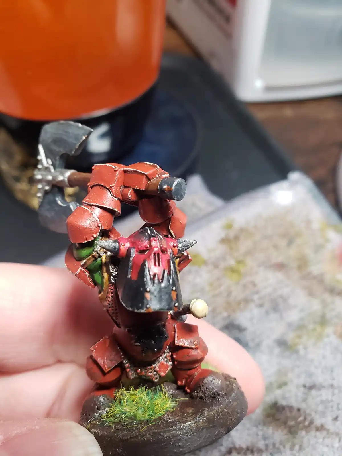 A hand holds a painted miniature of a red-armored, horned warrior with a black helmet and a large axe, standing on a muddy…