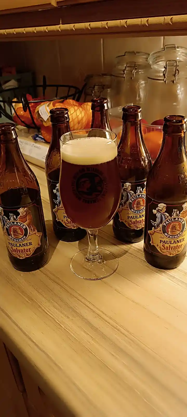 A glass of dark beer with a thick white head sits on a wooden surface surrounded by several Paulaner Salvator Doppelbock…