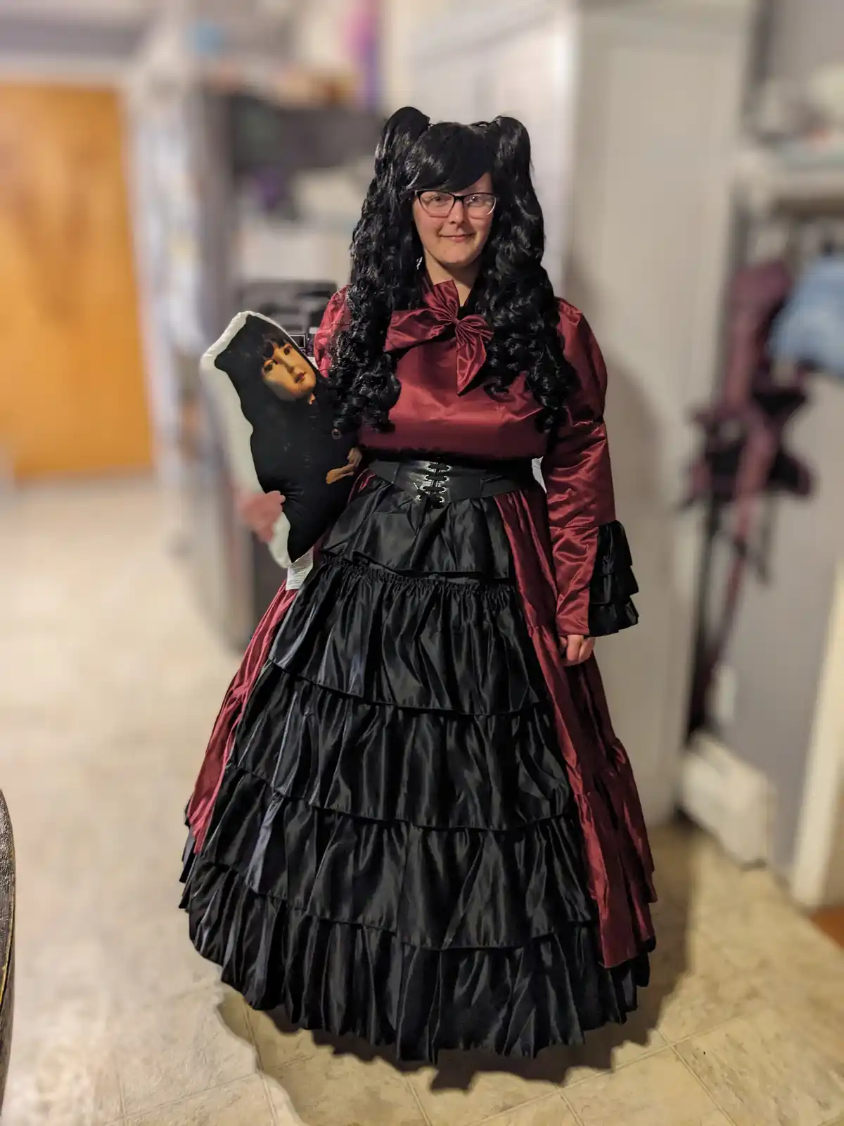 A person wearing a black and red Victorian-style dress with a corset and long black wig styled in twin buns, holding a large…