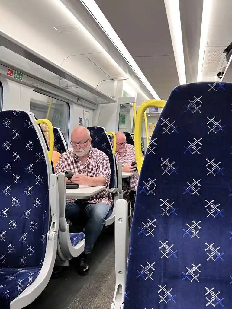 Inside a modern train carriage with blue patterned seats and yellow handrails, two identical-looking men in red-and-white…