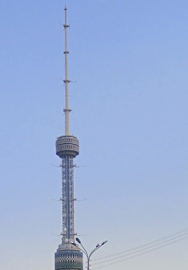 A tall, slender telecommunications tower with a circular observation deck near the top, set against a clear blue sky. The…