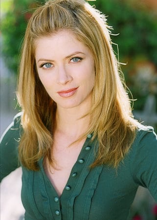 A woman with blonde hair and blue eyes, wearing a green button-up shirt, looks directly at the camera with a neutral…