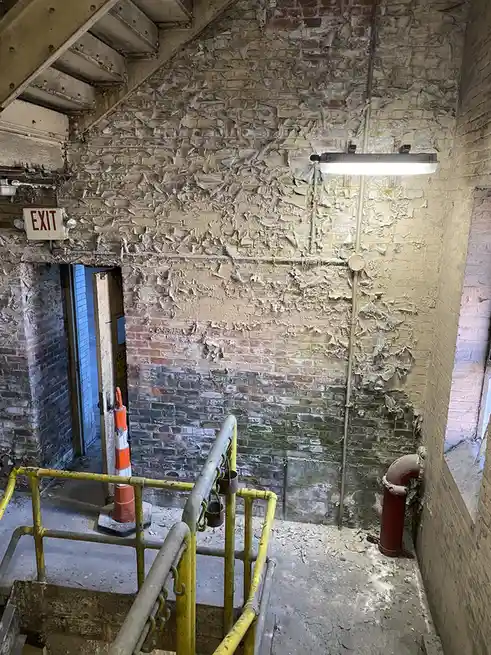 A dilapidated industrial stairwell with peeling paint on brick walls, a yellow railing, and an illuminated red 'EXIT' sign…