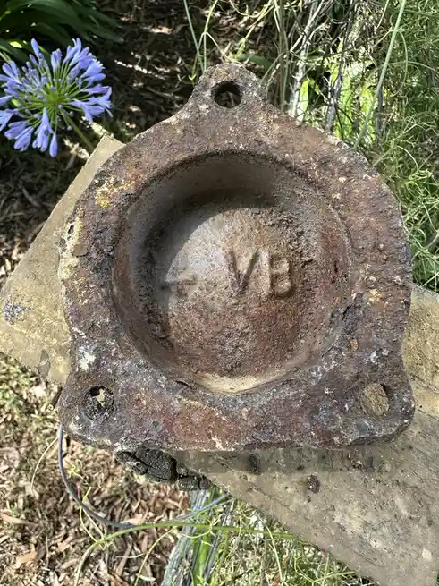 A rusty, weathered metal pipe fitting rests on a wooden surface outdoors, surrounded by greenery and a purple flower. The…