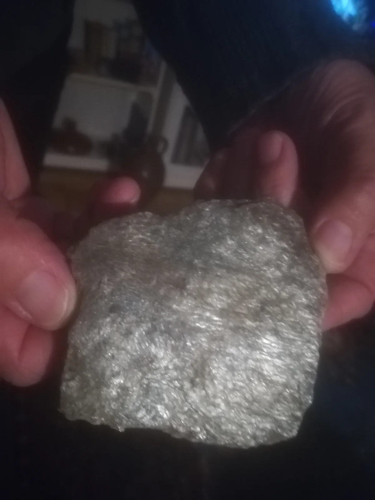 Two hands hold up a rough, grayish, crystalline rock with a glittery, uneven surface. The background is a dimly lit room…