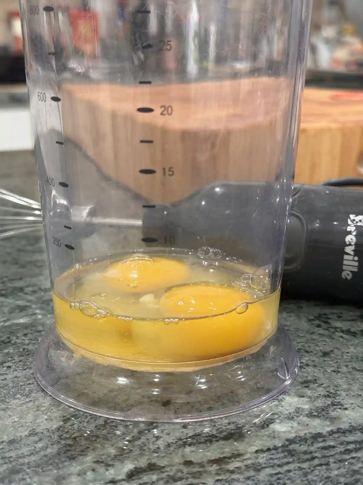 A clear plastic blender cup with measurement markings sits on a speckled countertop, containing two intact, unbroken egg…