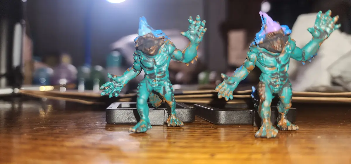 Two teal and brown painted monster figurines stand on a wooden surface, both wearing party hats—one blue, one purple—and…