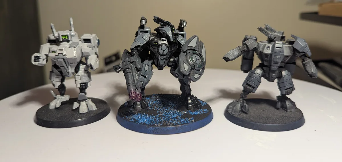 Three painted miniature mechs stand side-by-side on a white surface. The left mech is mostly white with green eyes. The…