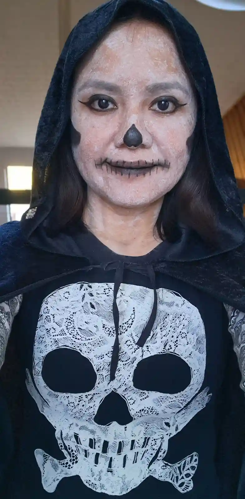 A person wearing a black hooded cloak over a black t-shirt with a white skull and crossbones graphic, has white face paint…