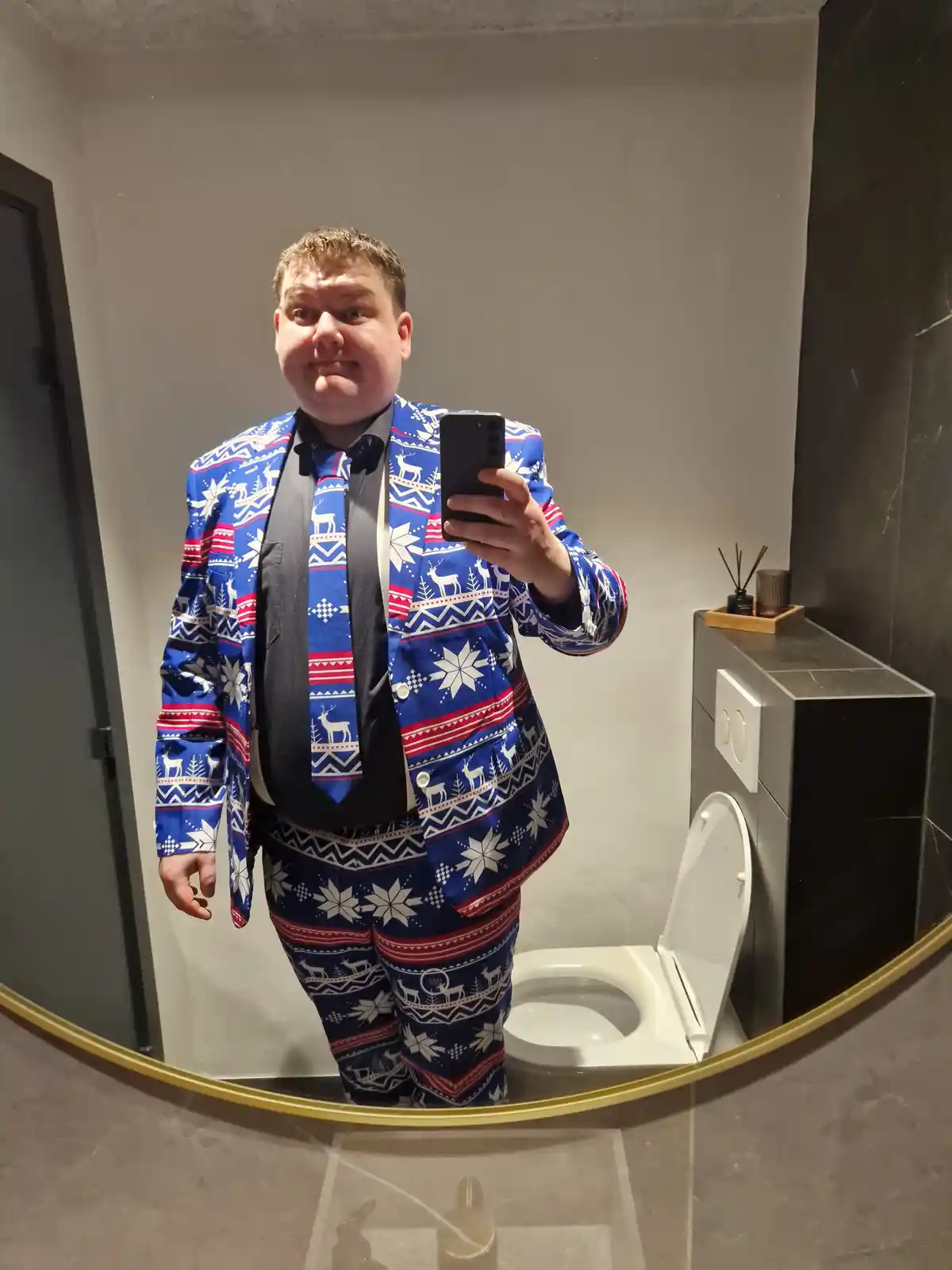 A man takes a mirror selfie in a bathroom while wearing a full blue, red, and white holiday-themed suit with reindeer and…
