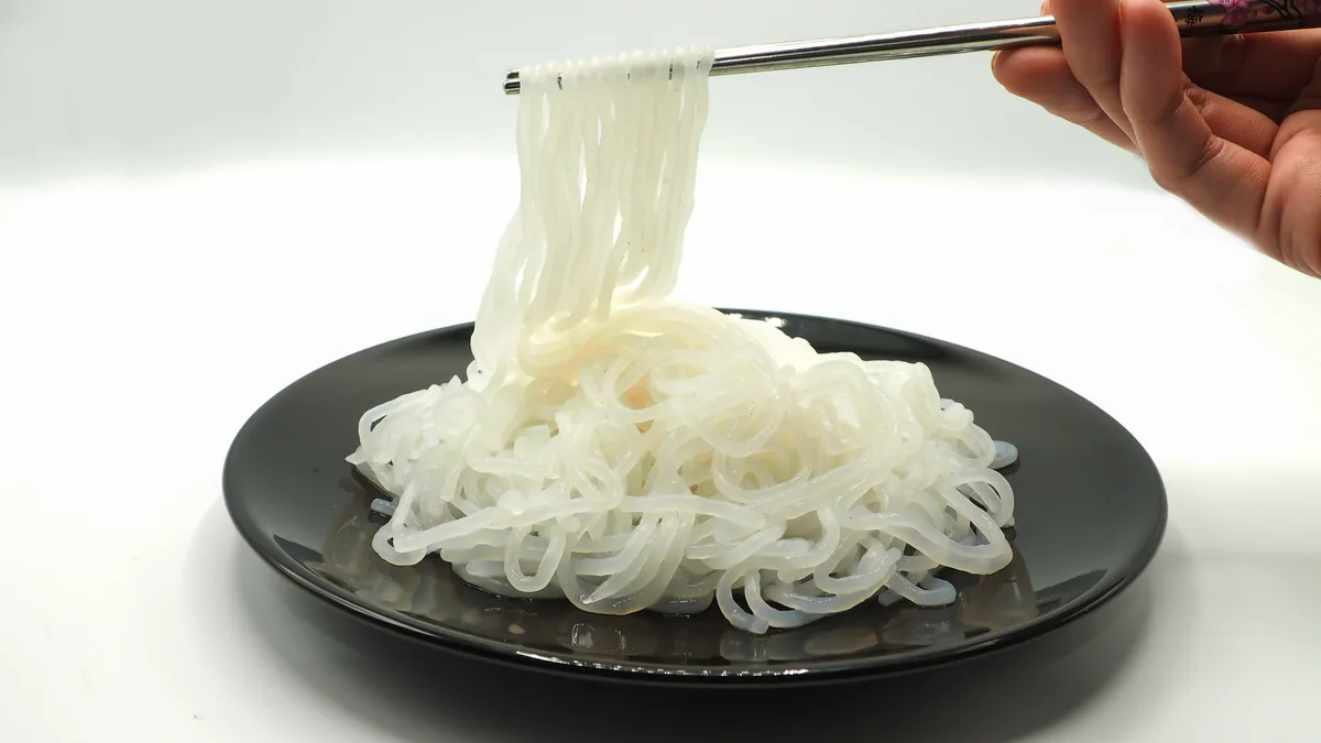 A hand uses chopsticks to lift a bundle of translucent, white rice noodles from a black plate against a white background…