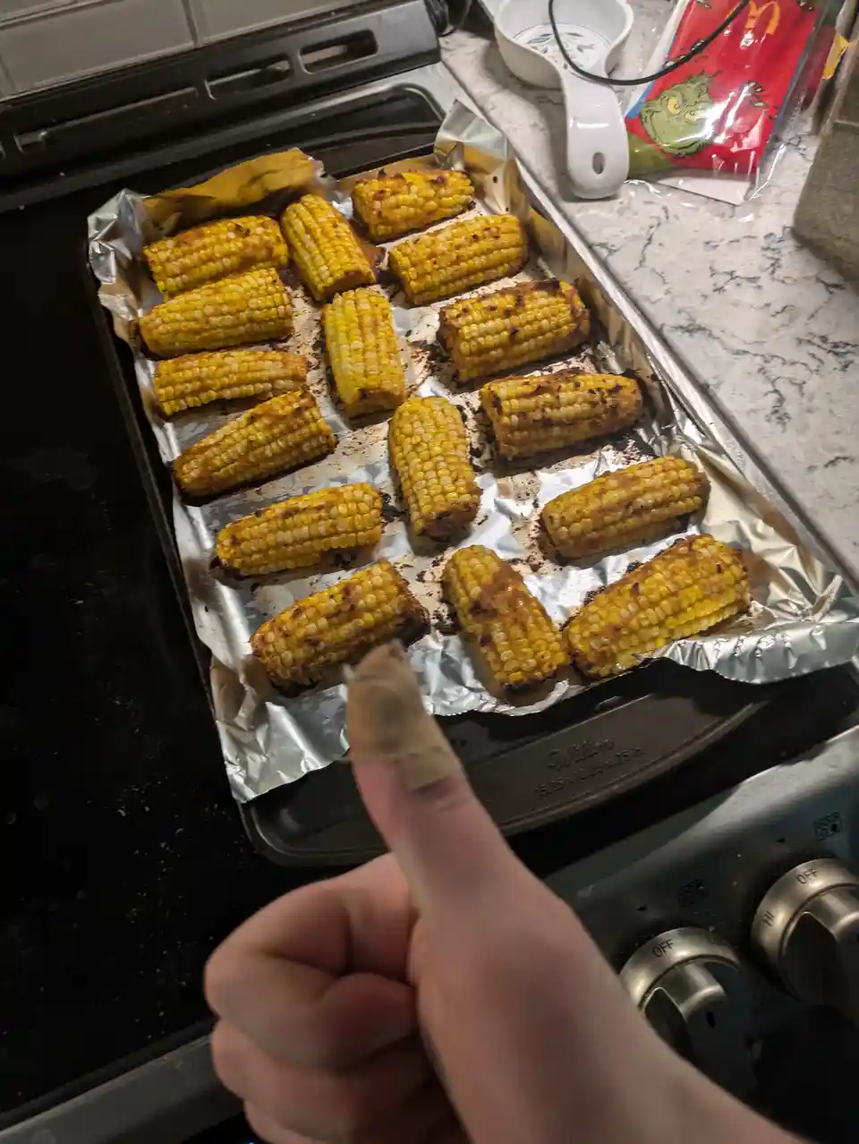 A hand with a bandaged thumb points at a baking tray lined with aluminum foil holding numerous pieces of roasted corn on the…