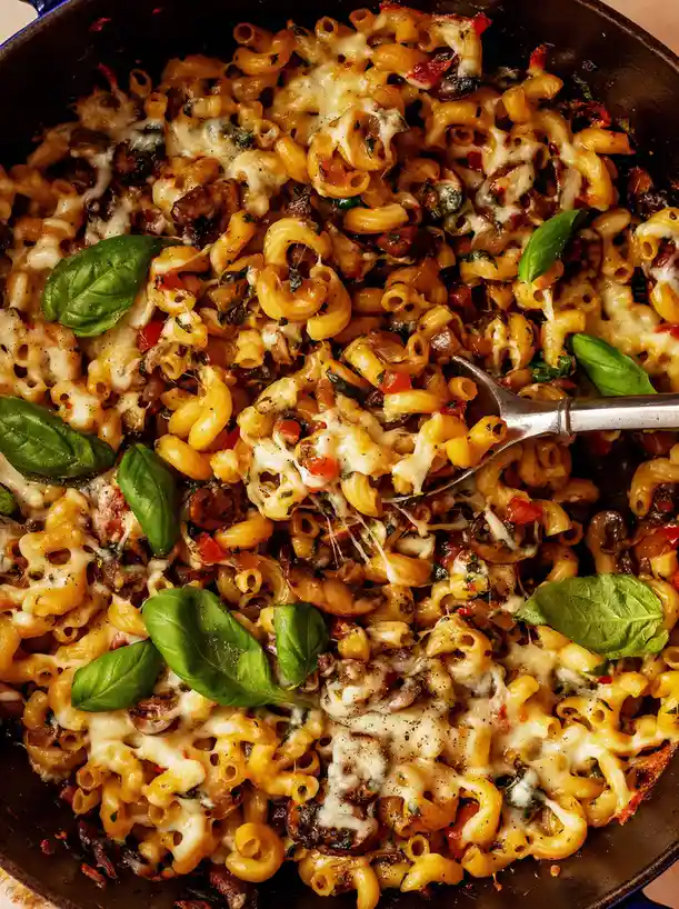 a close-up photo of a skillet filled with cheesy macaroni pasta mixed with mushrooms, tomatoes, and fresh basil leaves, with…