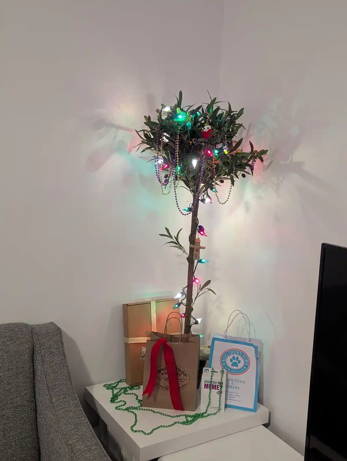 A potted olive tree is decorated with colorful Christmas lights, purple and gold beads, and small ornaments, standing on a…