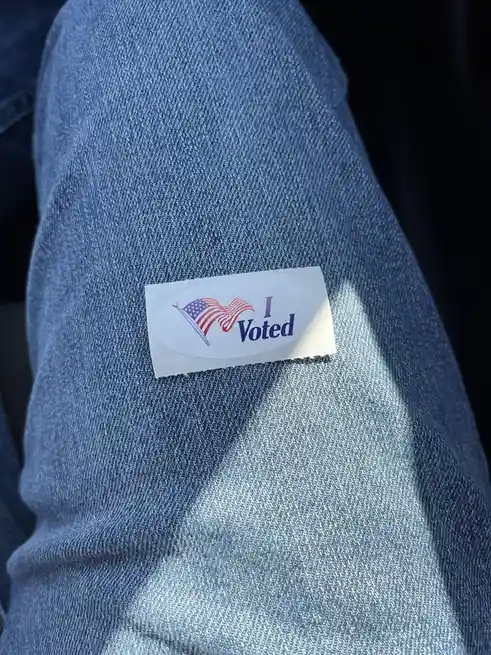 A close-up photo shows an 'I Voted' sticker, featuring a stylized American flag forming a heart shape, stuck on the denim…