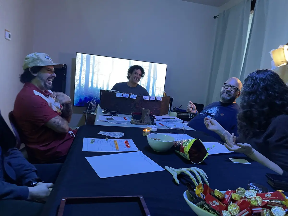 A group of people are gathered around a table covered in papers, dice, snacks, and a plastic skeleton hand, clearly engaged…