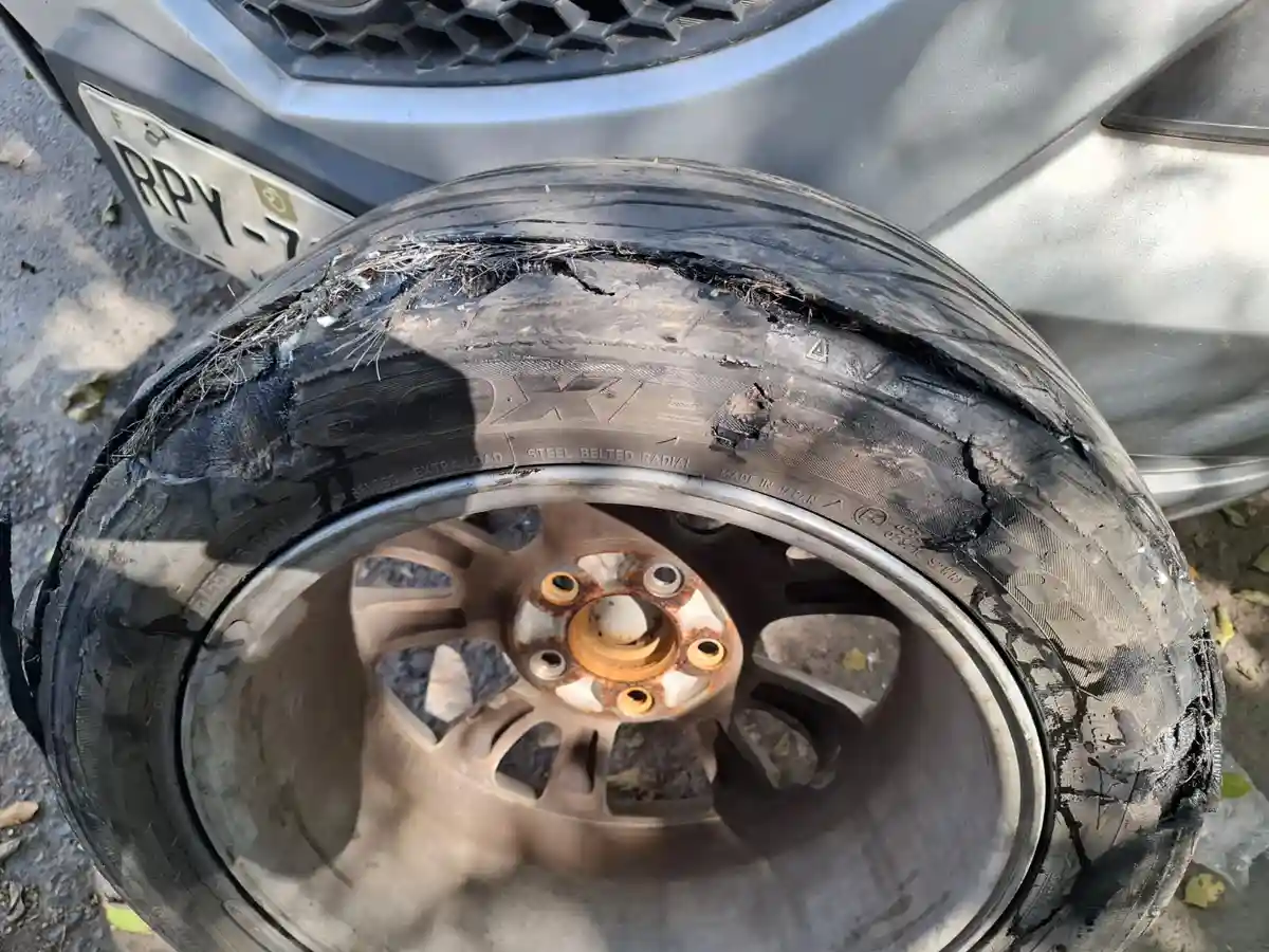A close-up photo shows a severely damaged car tire with torn rubber and embedded debris, resting on the ground near a silver…