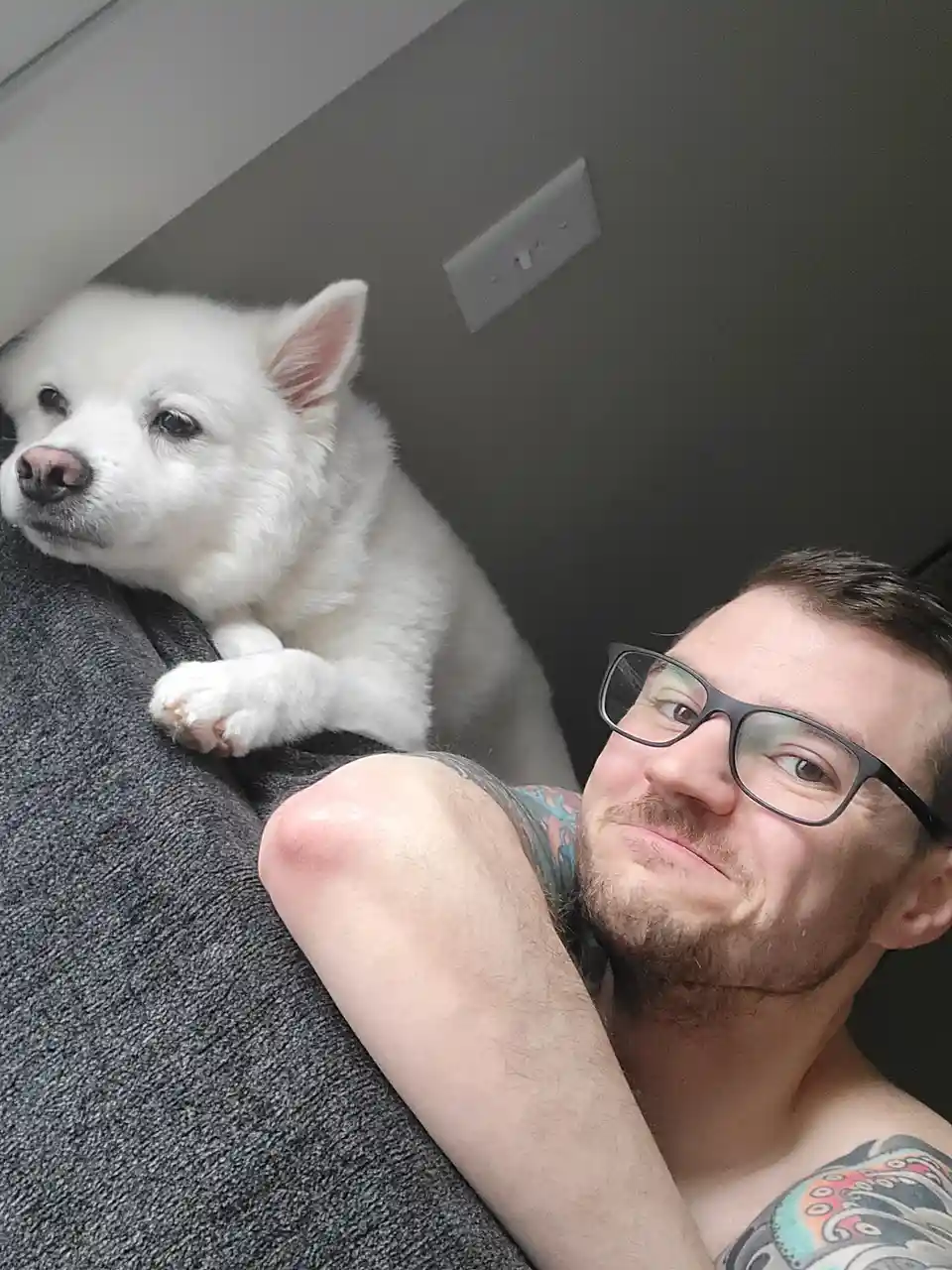 A man with glasses and tattoos takes a selfie while a fluffy white puppy rests on his shoulder, looking away. The man has a…