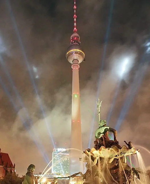 A nighttime photo of the Berlin TV Tower illuminated with red and white lights, with beams of light shooting from its top…