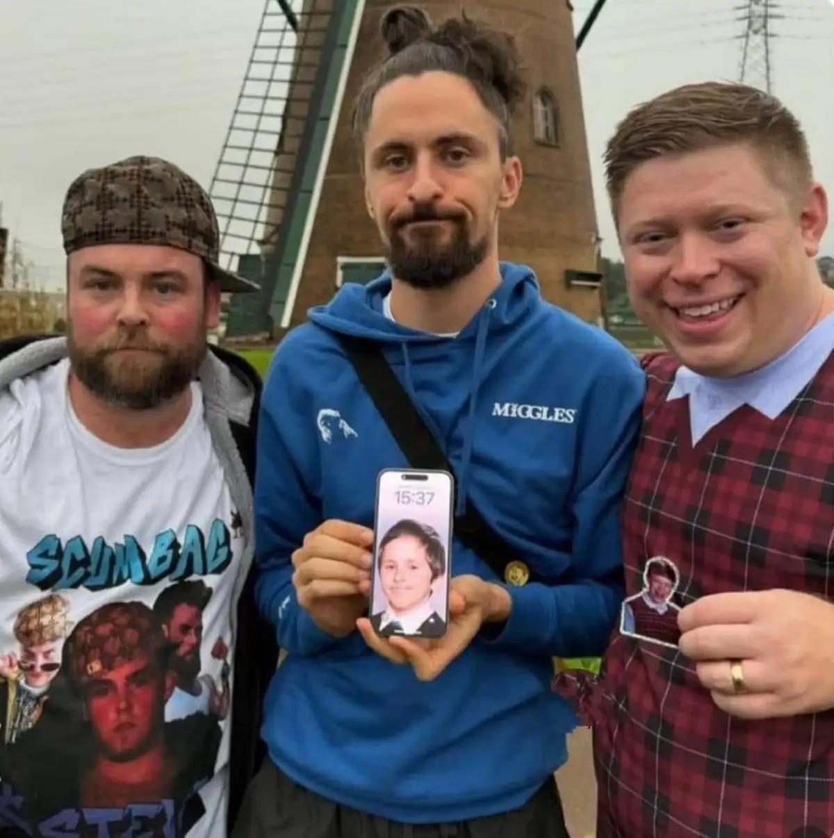 Three men stand outdoors in front of a windmill. The man in the center holds up a smartphone displaying a photo of a young…