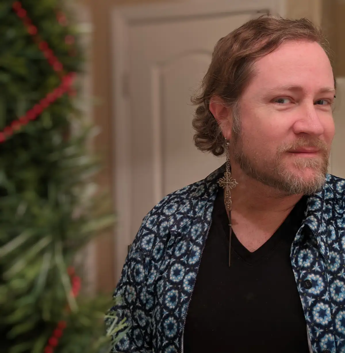 A man with shoulder-length brown hair and a beard, wearing a patterned blue shirt over a black v-neck, looks off to the side…