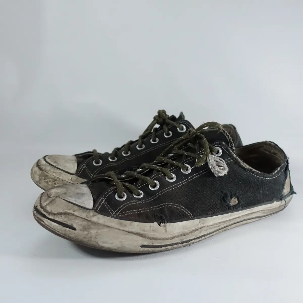 A pair of heavily worn, dirty black canvas sneakers with frayed laces and torn fabric, set against a plain white background…