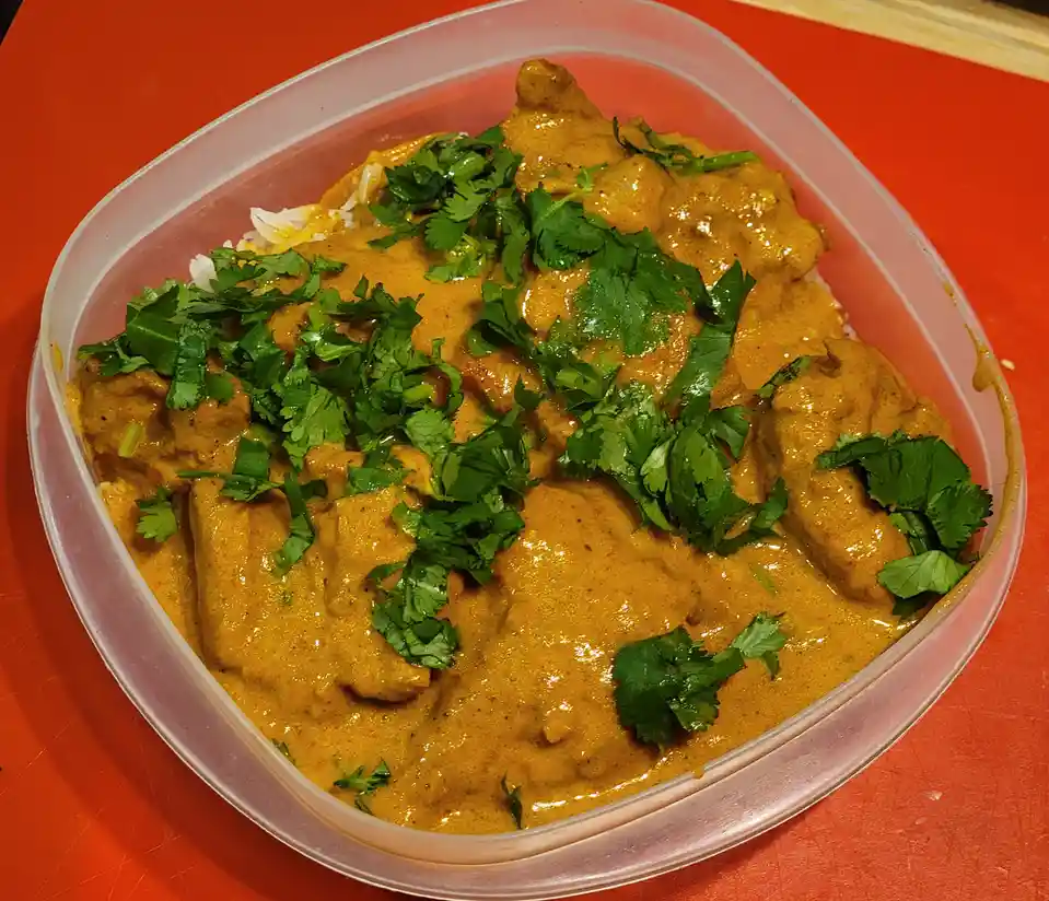 A close-up photo shows a clear plastic container filled with a creamy orange curry sauce over rice, generously topped with…