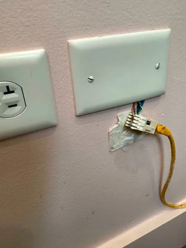 A close-up photo shows a yellow ethernet cable plugged directly into a wall outlet cover that has been ripped open, exposing…