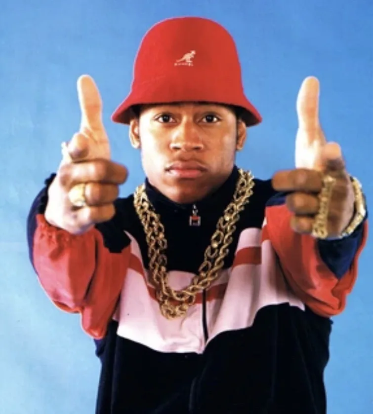 a young black man wearing a red Kangol bucket hat, a chunky gold chain, and a red, white, and black tracksuit, gives two…